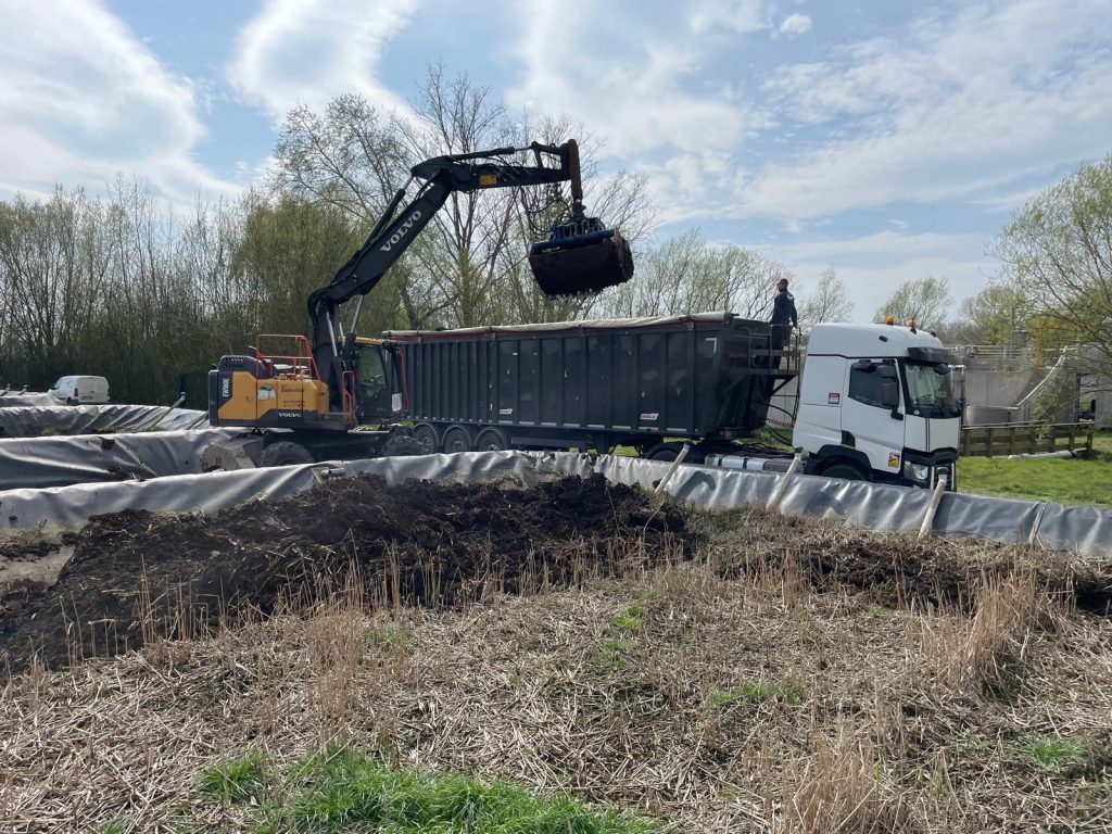 Pelle Volvo floquée Delestrez chargeant les sédiments d'un bassin d'assainissement dans une benne, ouvrage de traitement visible à l'arrière-plan